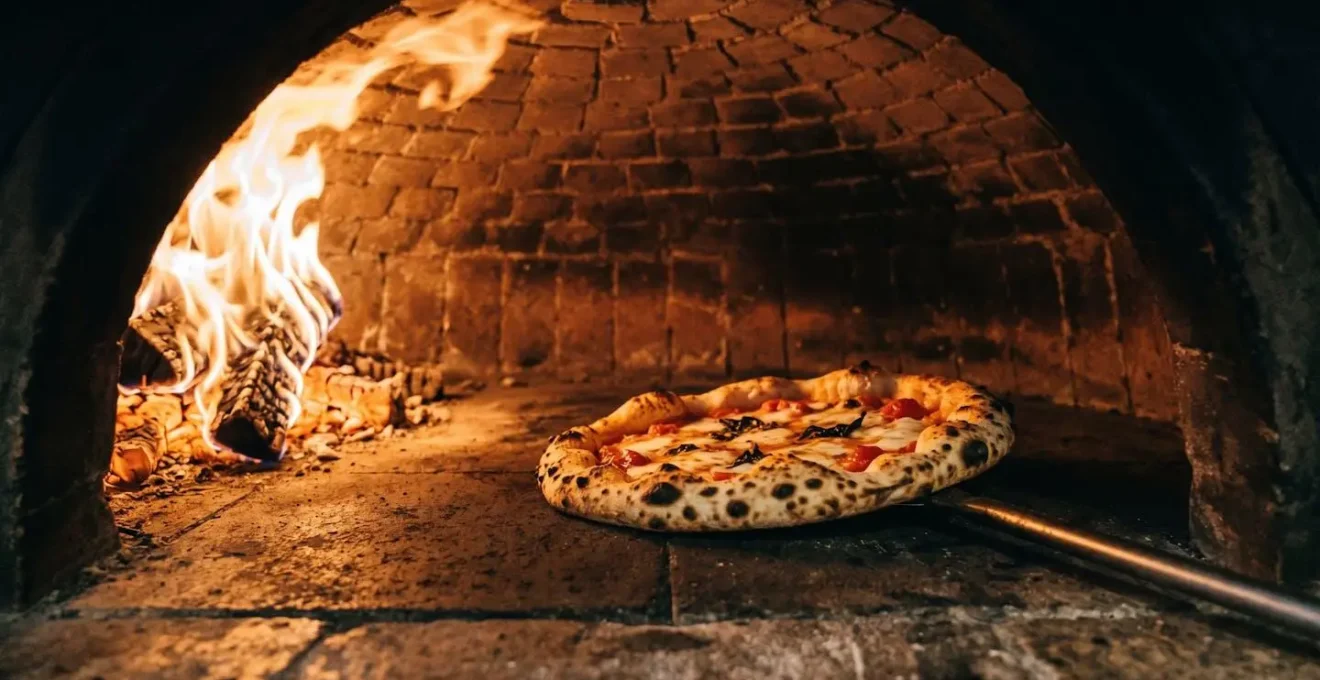 Intérieur d'un four à bois napolitain avec pizza en cuisson sur sole en pierre, flammes vives sur le côté et taches léopardées sur le bord de la pizza